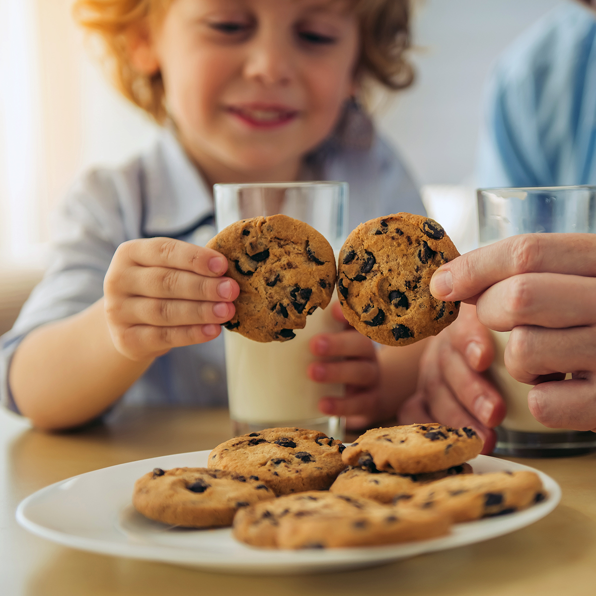 child cheersing cookies with another person over a plate of cookies and cups of milk
